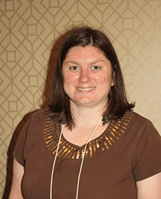 Casey Robertson smiles against a tan background, wearing shoulder-length, brown hair and a brown top.